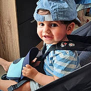 Lucas a rejoint le concours — aidez-le/la à gagner de superbes lots ! toddler, child, smiling, cap, blue_cap, striped_shirt, stroller, indoor, furniture, window, curtains, shelf, toy, happy, seated, person, face, baby, clothing, footwear
