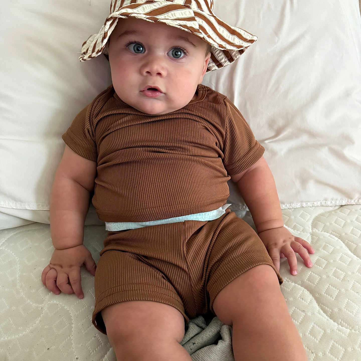 Liam Gabriel joined the competition — help win amazing prizes! baby, blue_eyes, brown_clothing, casual, child, chubby_cheeks, curious, cute, fabric, hat, indoors, infant, light, mattress, pillow, portrait, sitting, soft, striped_hat, young_child