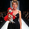 woman, dog, dress, red_hat, runway, fashion, audience, black_dress, pet, cute, portrait, smile, event, indoor, leash, small_dog, model, holding, catwalk, lighting