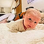 baby, infant, newborn, tummy_time, blanket, bed, soft_texture, blue_eyes, face, cheeks, onesie, lying_down, portrait, indoor, home_interior, pillow, curious_expression, close_up, adorable, nursery