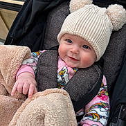 Love is registered to the contest to win money with this photo: baby, child, smiling, hat, knit_hat, pom_poms, blanket, stroller, infant, cute, cozy, unicorn_pattern, colorful_clothing, pink, face, hand, warm_clothing, indoor, portrait, happy