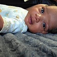 Klins is registered to the contest to win money with this photo: baby, infant, child, portrait, lying_down, blanket, indoor, close_up, face, eyes, smiling, curious, short_hair, dark_hair, shirt, patterned_clothing, soft_texture, cozy, head, photography