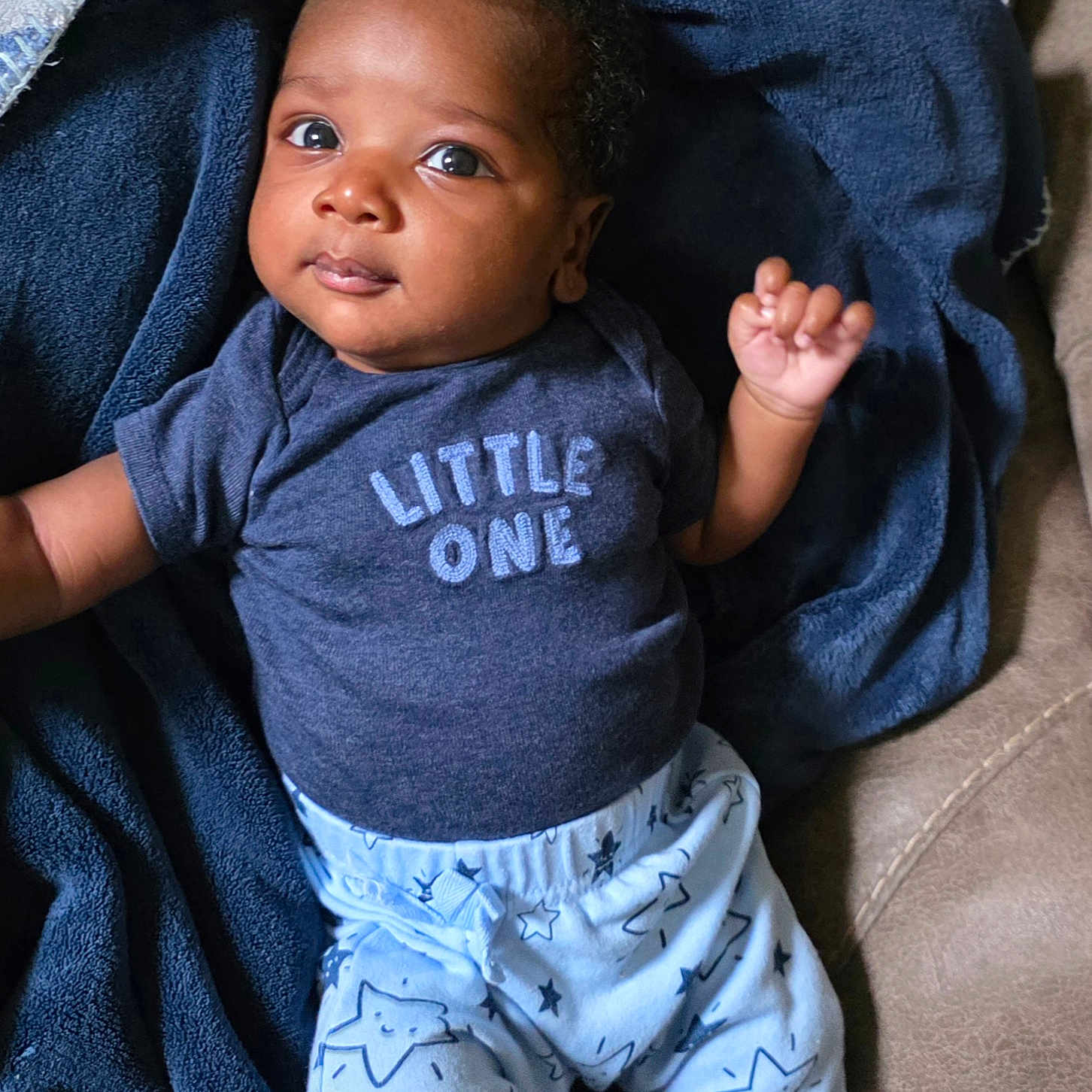 Klins is registered to the contest to win money with this photo: baby, infant, child, portrait, smiling, wide_eyed, lying_down, blanket, navy_blue, clothing, onesie, shirt, pants, star_pattern, couch, indoor, cozy, raised_hand, dark_skin, close_up