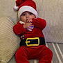 baby, infant, santa_hat, red_clothing, holiday_costume, couch, pillows, cozy, indoors, child, cute, festive, sitting, plush, face, hand, feet, striped_blanket, soft_furniture, wide_eyes