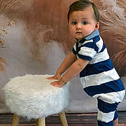 Maylon a rejoint le concours — aidez-le/la à gagner de superbes lots ! toddler, child, blue_and_white_striped_clothing, striped_romper, standing, white_furry_stool, wooden_stool_legs, indoor, studio, brown_backdrop, dried_plants, curious_expression, sandals, bear_face_sandals, flooring, portrait, baby, hands_on_stool, short_hair, bracelet