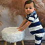 toddler, child, blue_and_white_striped_clothing, striped_romper, standing, white_furry_stool, wooden_stool_legs, indoor, studio, brown_backdrop, dried_plants, curious_expression, sandals, bear_face_sandals, flooring, portrait, baby, hands_on_stool, short_hair, bracelet