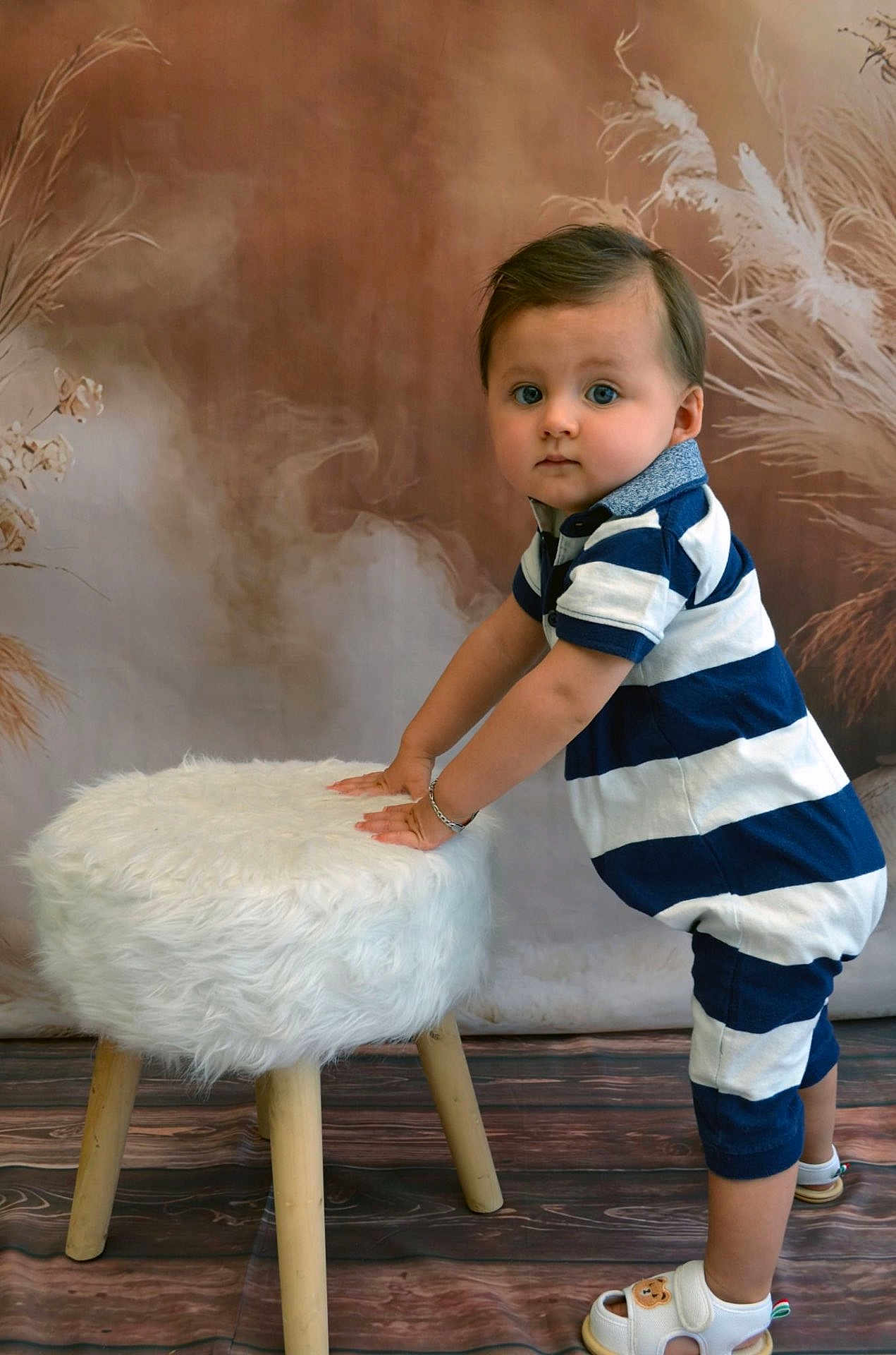 Maylon a rejoint le concours — aidez-le/la à gagner de superbes lots ! toddler, child, blue_and_white_striped_clothing, striped_romper, standing, white_furry_stool, wooden_stool_legs, indoor, studio, brown_backdrop, dried_plants, curious_expression, sandals, bear_face_sandals, flooring, portrait, baby, hands_on_stool, short_hair, bracelet