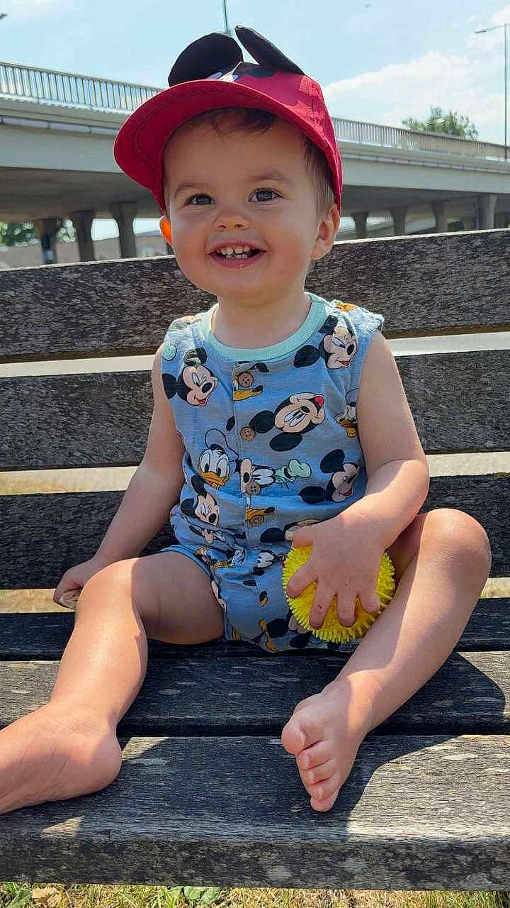 Lorenzo a rejoint le concours — aidez-le/la à gagner de superbes lots ! toddler, child, smiling, outdoor, bench, cap, red_cap, mickey_mouse, donald_duck, onesie, toy, yellow_ball, happy, sunny, daylight, grass, person, barefoot, cute, playful