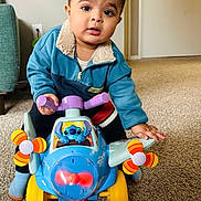 Hussain joined the competition — help win amazing prizes! toddler, child, toy_airplane, blue_jacket, indoor, carpet, propeller, yellow_wheels, orange_wheel, blue_toy, person, play, cute, home, curious, floor, furniture, door, expression, hand