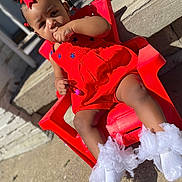 Maliha joined the competition — help win amazing prizes! toddler, child, red_dress, red_chair, white_socks, outdoor, sunlight, cute, baby, sitting, thumb_sucking, hair_ribbons, beads, plastic_chair, concrete, shadow, curious_expression, footwear, portrait, young_child