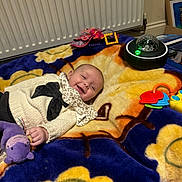 Emmylou is registered to the contest to win money with this photo: baby, blanket, smiling, toy, stuffed_animal, indoor, radiator, colorful, playtime, child, clothing, purple, yellow, orange, blue, black, plastic, happy, person, floor