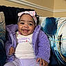 baby, child, smile, purple_jacket, headband, bow, couch, cushion, happy, indoor, person, clothing, seated, cute, infant, portrait, cozy, homedecor, warm, playful