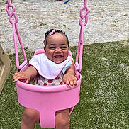 Brazille is registered to the contest to win money with this photo: baby, child, swing, playground, pink, smile, happy, grass, outdoor, bib, hair_clips, footwear, fence, toy, sunny, toddler, seat, joyful, person, summer