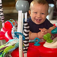 Isaiah joined the competition — help win amazing prizes! baby, child, smiling, happy, indoors, toys, activity_center, play, colorful, red, blue, green, fabric, pattern, clothing, hands, face, person, furniture, living_room