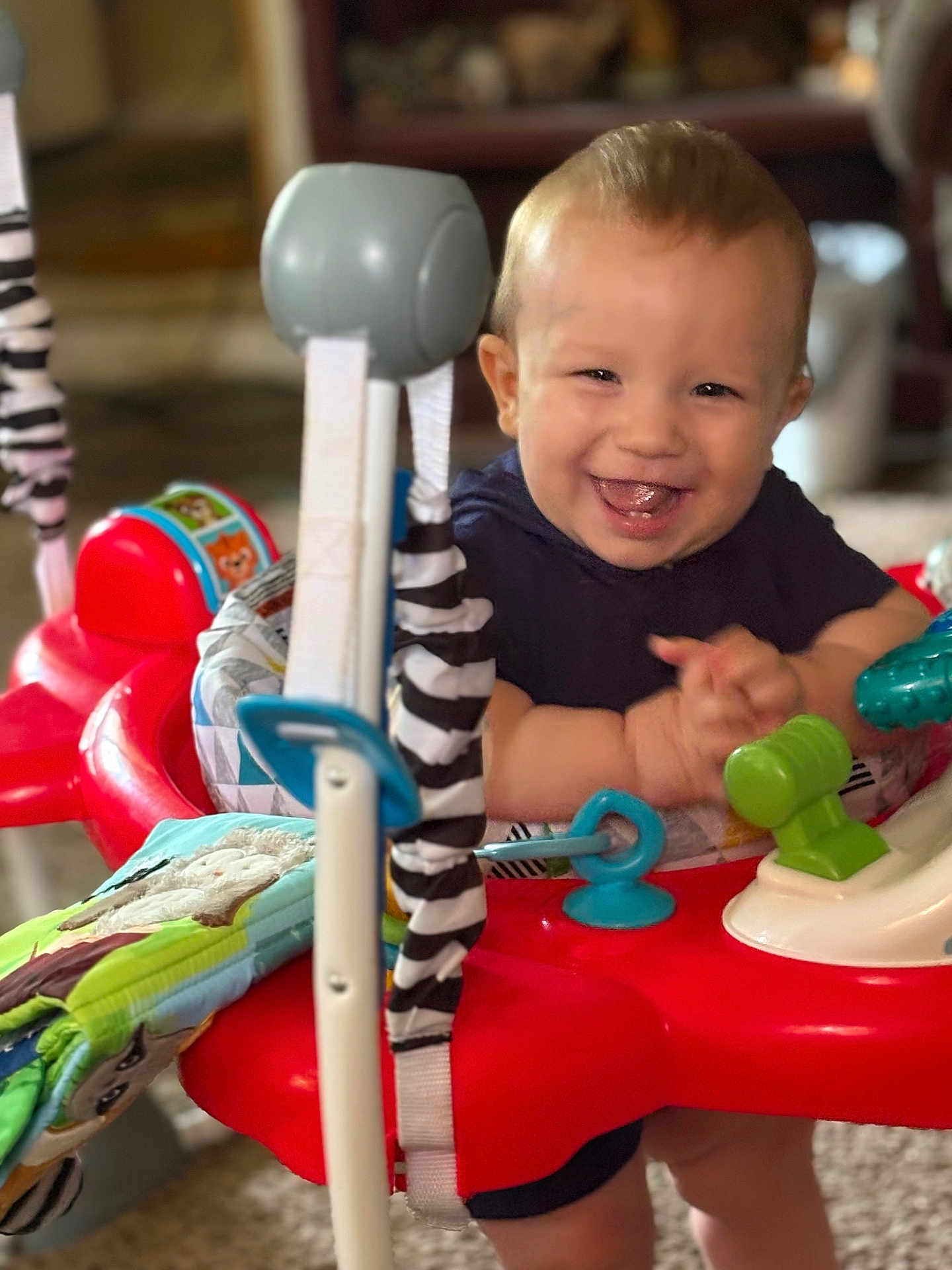 Isaiah joined the competition — help win amazing prizes! baby, child, smiling, happy, indoors, toys, activity_center, play, colorful, red, blue, green, fabric, pattern, clothing, hands, face, person, furniture, living_room