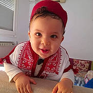Iyed a rejoint le concours — aidez-le/la à gagner de superbes lots ! baby, child, smiling, face, traditional_clothing, red_hat, indoor, couch, pillow, curly_hair, hand, white_wall, window, radiator, portrait, happy, cute, person, home, infant