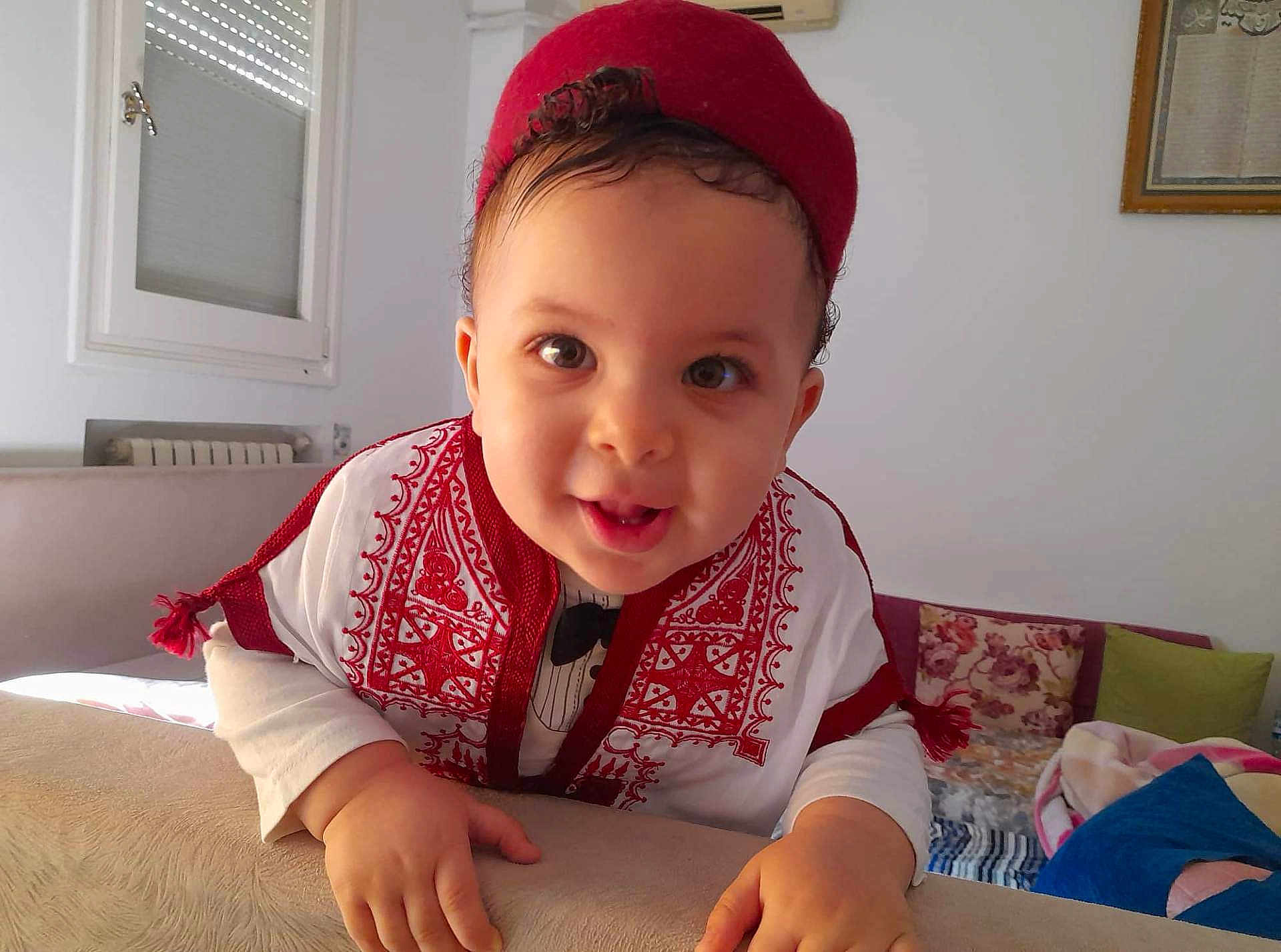 Iyed a rejoint le concours — aidez-le/la à gagner de superbes lots ! baby, child, smiling, face, traditional_clothing, red_hat, indoor, couch, pillow, curly_hair, hand, white_wall, window, radiator, portrait, happy, cute, person, home, infant