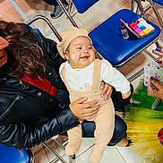 Ethan is registered to the contest to win money with this photo: baby, child, smiling, happy, infant, portrait, indoor, seated, held, adult, woman, hat, beanie, overalls, white_shirt, chairs, makeup, cosmetics, tile_floor, family