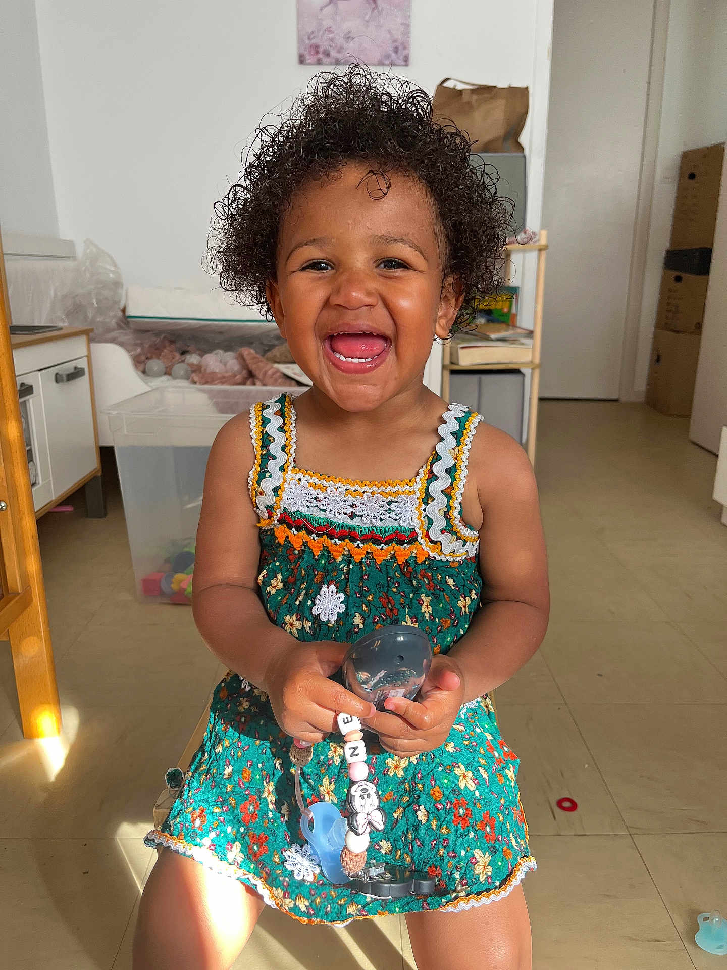 Nejma a rejoint le concours — aidez-le/la à gagner de superbes lots ! box, cabinet, child, curly_hair, cute, dress, floor, furniture, happy, indoor, pacifier, person, playful, portrait, room, shadow, smiling, sunlight, toddler, toy