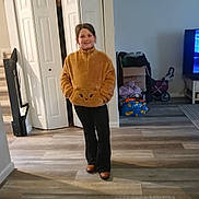 Peyton is registered to the contest to win money with this photo: black_pants, brown_pullover, brown_shoes, casual_pose, child, clothing, cozy, fuzzy_pullover, home, household_items, indoor, living_room, open_closet, person, portrait, smile, standing, tv, wooden_floor, young_person