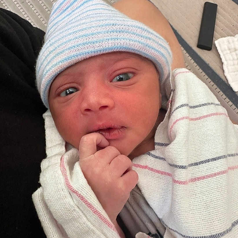 Noel is registered to the contest to win money with this photo: newborn, baby, infant, wrapped, blanket, hat, striped_hat, hospital, person, hand, face, eyes, indoor, carpet, floor, resting, cute, small, cozy, portrait