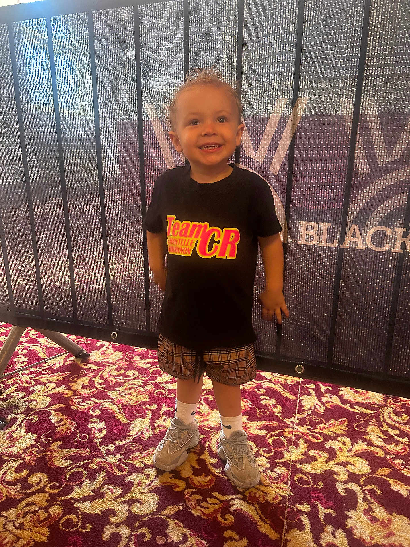 Kevin joined the competition — help win amazing prizes! toddler, child, smiling, standing, carpet, patterned_carpet, black_tshirt, text_on_tshirt, plaid_shorts, sneakers, white_socks, indoor, mesh_barrier, event, person, happy, curly_hair, footwear, casual_clothing, background_text
