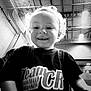 Kevin is registered to the contest to win money with this photo: child, toddler, smiling, curly_hair, indoor, gymnasium, ceiling, lighting, black_and_white, portrait, person, shirt, text_on_shirt, happy, young_child, head, face, clothing, background, sports_facility