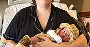 Christopher is registered to the contest to win money with this photo: adult, baby, newborn, parent, hospital, hospital_room, bed, portrait, family, holding, cradling, crocheted_clothing, knitted_hat, blanket, glasses, smiling, black_clothing, wooden_cabinets, medical_setting, birth
