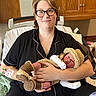 adult, baby, newborn, parent, hospital, hospital_room, bed, portrait, family, holding, cradling, crocheted_clothing, knitted_hat, blanket, glasses, smiling, black_clothing, wooden_cabinets, medical_setting, birth