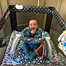 Antonio is registered to the contest to win money with this photo: baby, child, playpen, toy, blue_onesie, smiling, indoor, cushion, fabric, pattern, infant, happy, sitting, colorful, soft_toy, home, person, furniture, baby_toy, play_area