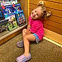child, smiling, sitting, carpet, wooden_wall, book_display, books, reading, colorful, indoor, young_girl, ponytails, pink_shirt, shorts, footwear, happy, portrait, playful, casual, floor
