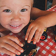 Maisie is registered to the contest to win money with this photo: child, smiling, toy, monster_truck, tablecloth, red, white, hands, fingernails, closeup, face, play, happy, indoor, person, cute, expression, childhood, fun, young