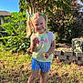 child, girl, smiling, outdoor, grass, tree, sunlight, pigtails, yellow_tshirt, striped_shorts, crocs, toy, pacifier, greenery, nature, happy, summer, playful, person, portrait
