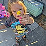 child, flower, yellow_flower, pink_nails, outdoor, sunlight, playing, toy, boots, concrete, smiling, tshirt, blonde_hair, person, sidewalk, fence, hand, happy, nature, daylight