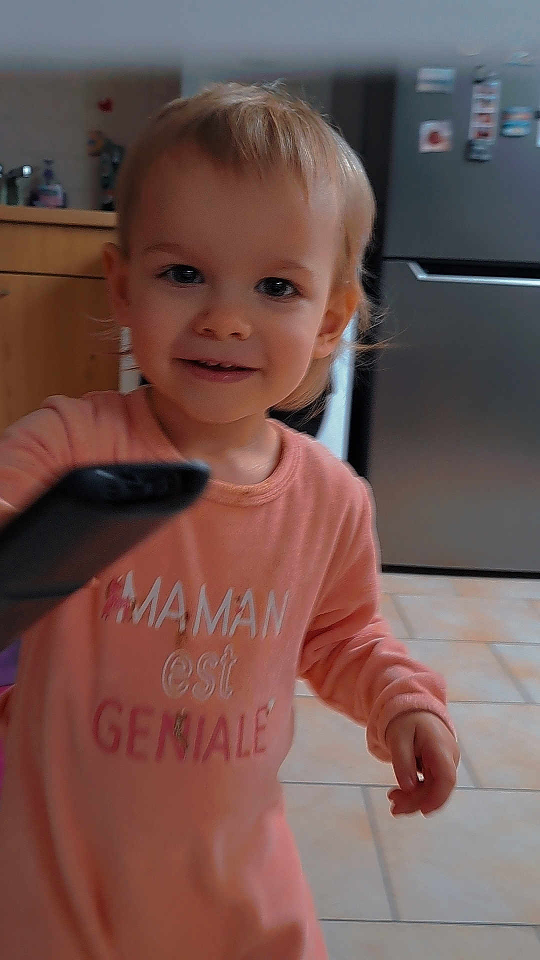 Laurianna a rejoint le concours — aidez-le/la à gagner de superbes lots ! blond_hair, cabinet, casual_clothing, child, curious, cute, face, floor_tiles, home, indoor, kitchen, person, pink_shirt, portrait, refrigerator, remote_control, small_hand, smile, toddler, young_child