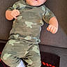 baby, child, couch, camouflage_clothing, pajamas, happy, smiling, white_socks, blanket, red, black, plaid_pattern, hair, face, person, infant, indoor, cute, joyful, relaxed