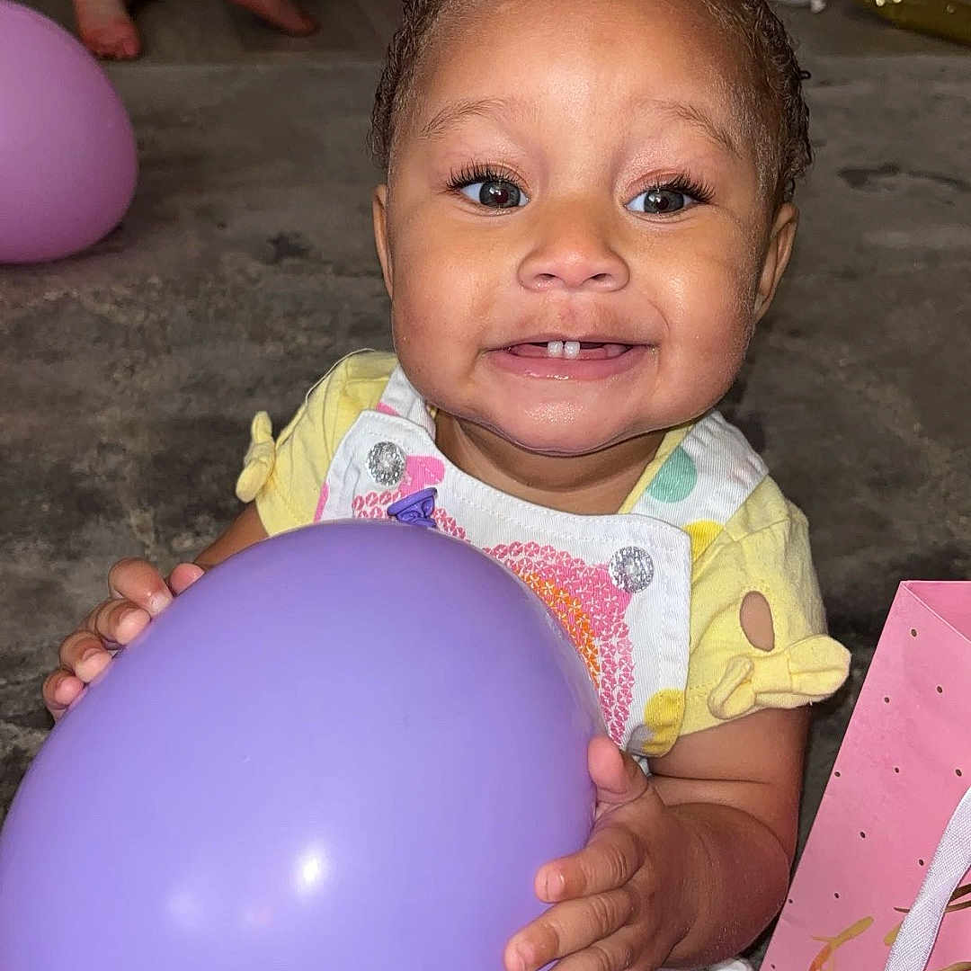 Madison is registered to the contest to win money with this photo: toddler, child, balloon, purple_balloon, smile, happy, indoors, carpet, party, birthday, playful, curly_hair, yellow_clothing, pink_gift_bag, background_child, floor, candid, cute, fun, celebration