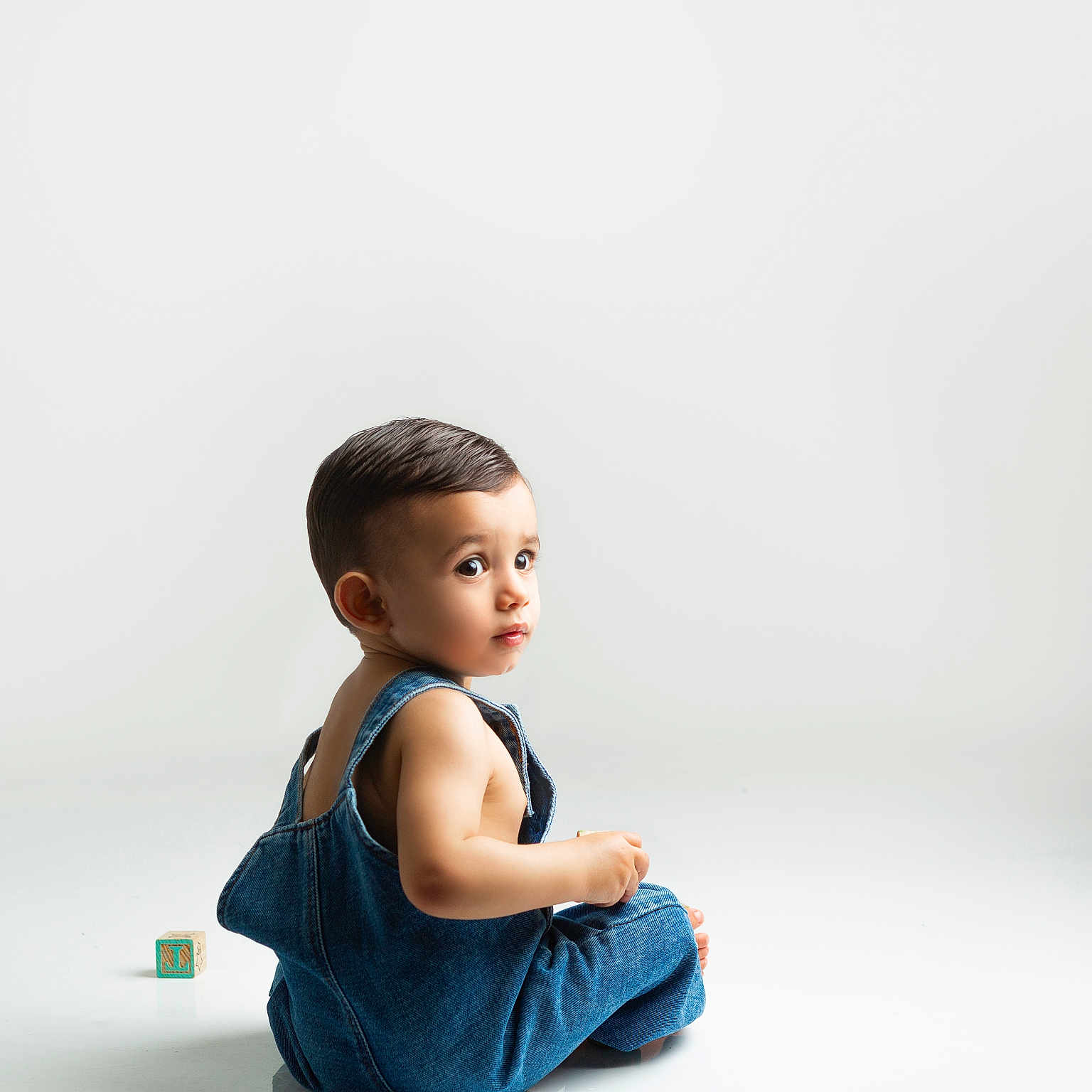 Ilhan joined the competition — help win amazing prizes! baby, casual_clothing, child, childhood, curious, cute, denim_overalls, floor, indoor, innocence, looking_back, minimalist, playtime, portrait, sitting, studio, toddler, toy_block, white_background, young_child