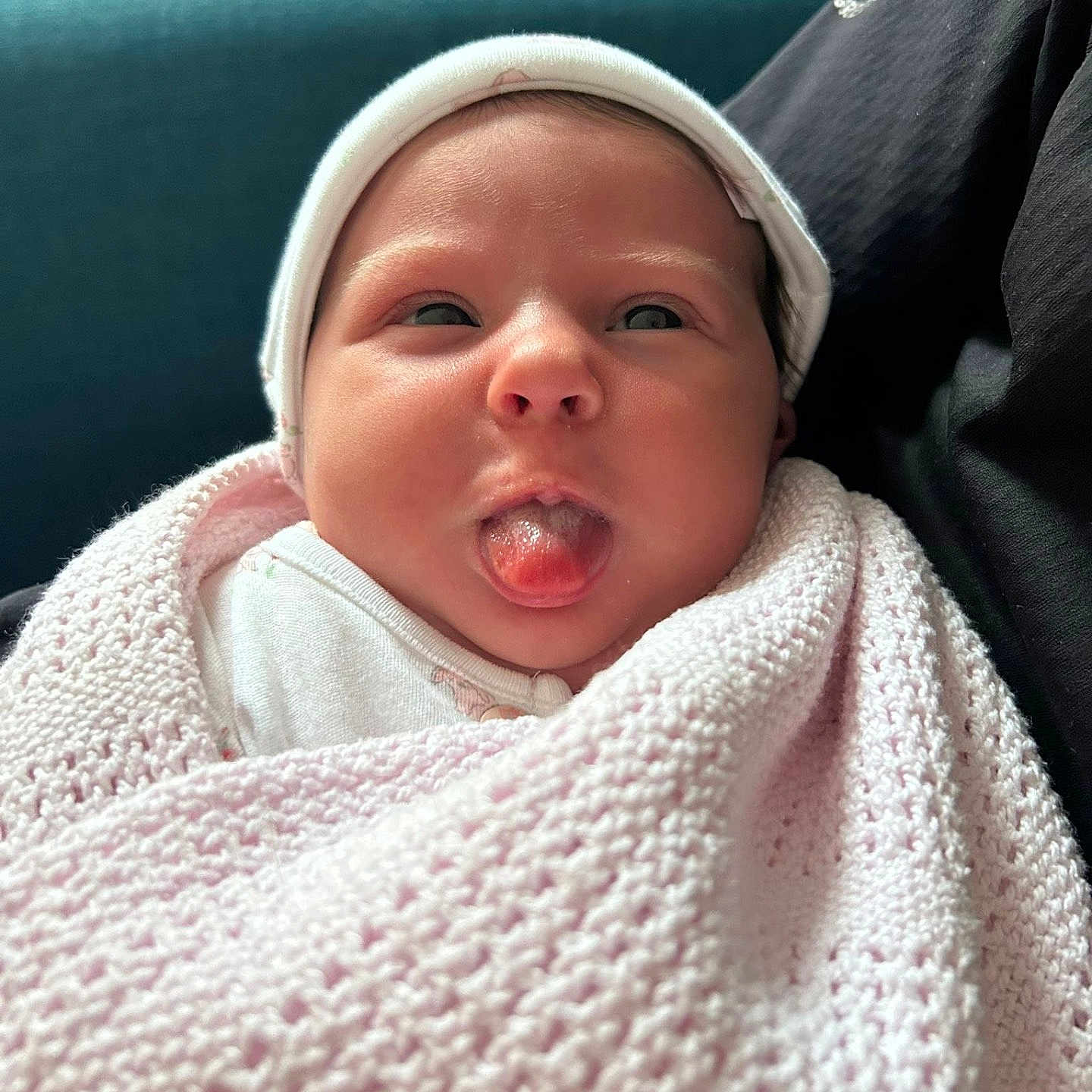 Maeve joined the competition — help win amazing prizes! baby, blanket, candid, child, cozy, cute, expression, face, hat, headwear, indoors, infant, knitwear, newborn, person, seated, skin, smile, tongue_out, wrapped
