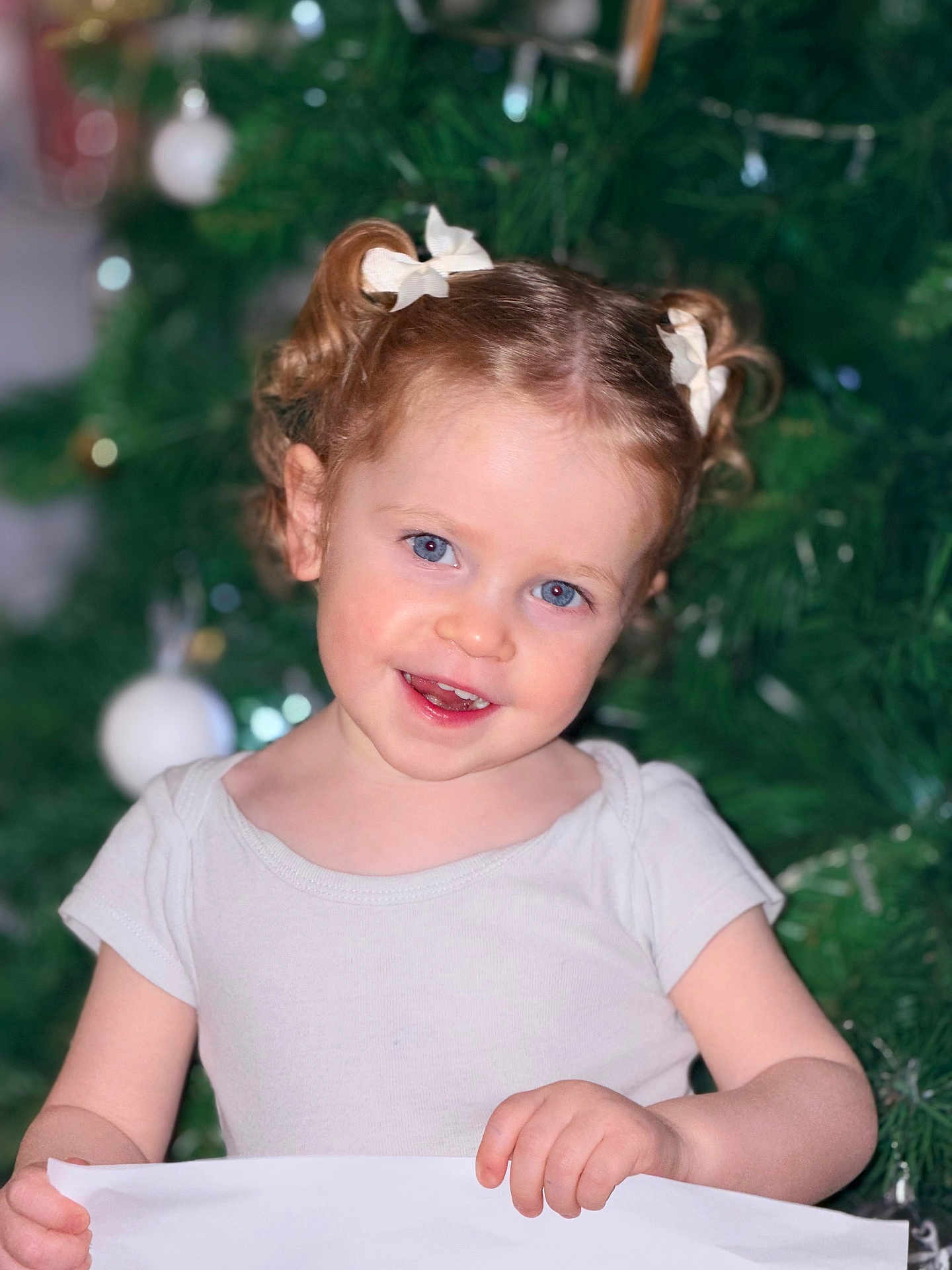 Rosalia participe au concours pour gagner de l'argent avec cette photo : child, toddler, smile, curly_hair, hair_bows, white_shirt, christmas_tree, ornaments, holiday, festive, portrait, blue_eyes, cute, indoors, holding_paper, happy, seasonal, decoration, celebration, person
