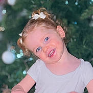 Rosalia participe au concours pour gagner de l'argent avec cette photo : child, toddler, smiling, curly_hair, blue_eyes, white_bow, white_shirt, portrait, christmas_tree, ornaments, holiday, indoors, hands, face, cute, happy, festive, background_blur, person, seasonal