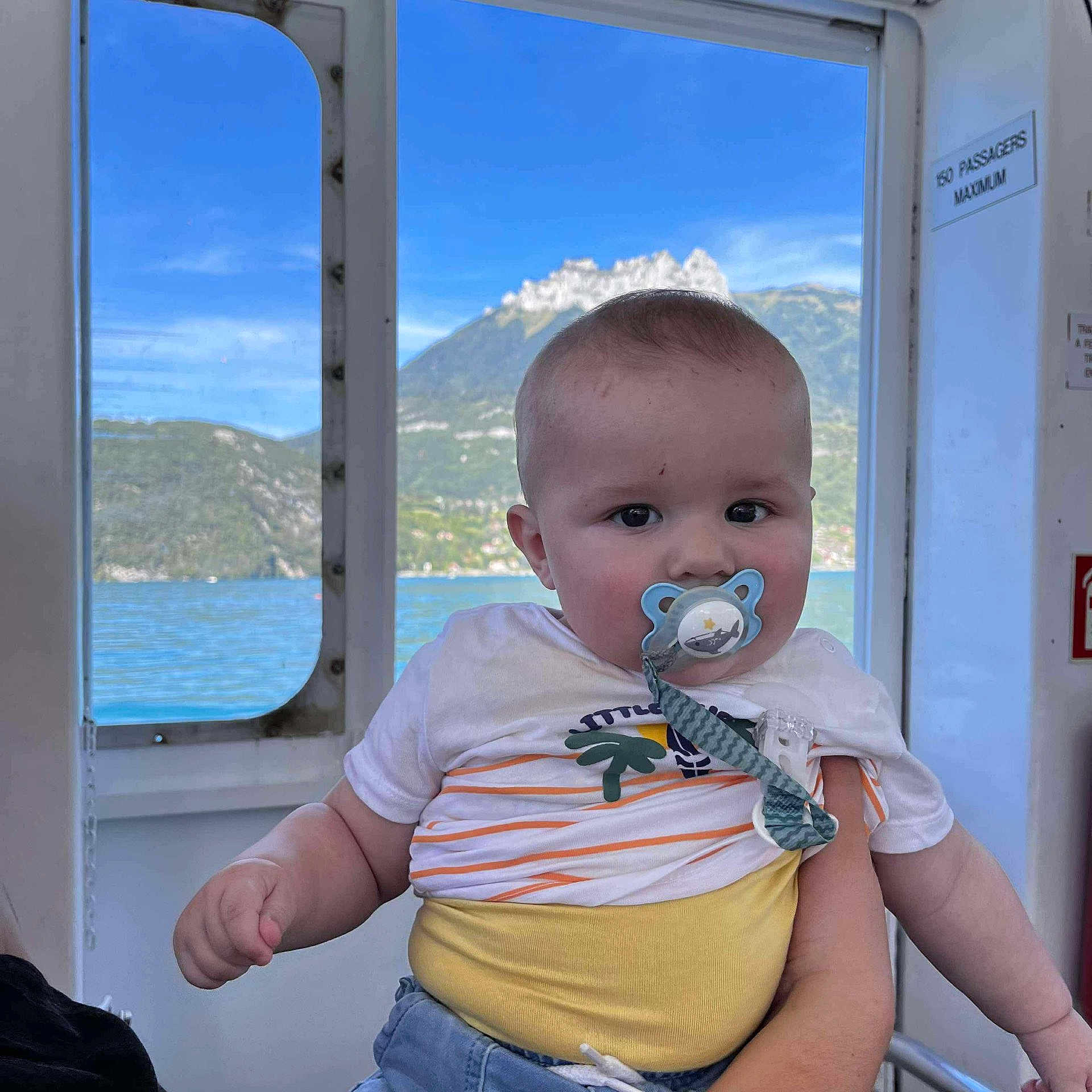 Lewis a rejoint le concours — aidez-le/la à gagner de superbes lots ! baby, pacifier, child, indoor, window, mountain, lake, water, blue_sky, person, holding, clothing, shirt, shorts, arm, hand, nature, scenic_view, transportation, boat