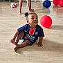 toddler, child, indoor, floor, balloons, toy, sports_jersey, paris_saint_germain, curious, sitting, barefoot, person, wooden_floor, red_balloon, blue_balloon, white_shorts, legs, home, playful, casual_clothing