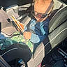 toddler, child, car_seat, sunglasses, curly_hair, chopsticks, blue_shirt, camouflage_pants, smile, sunlight, shadow, vehicle_interior, seatbelt, playful, casual_clothing, young_child, car, seat, portrait, happy
