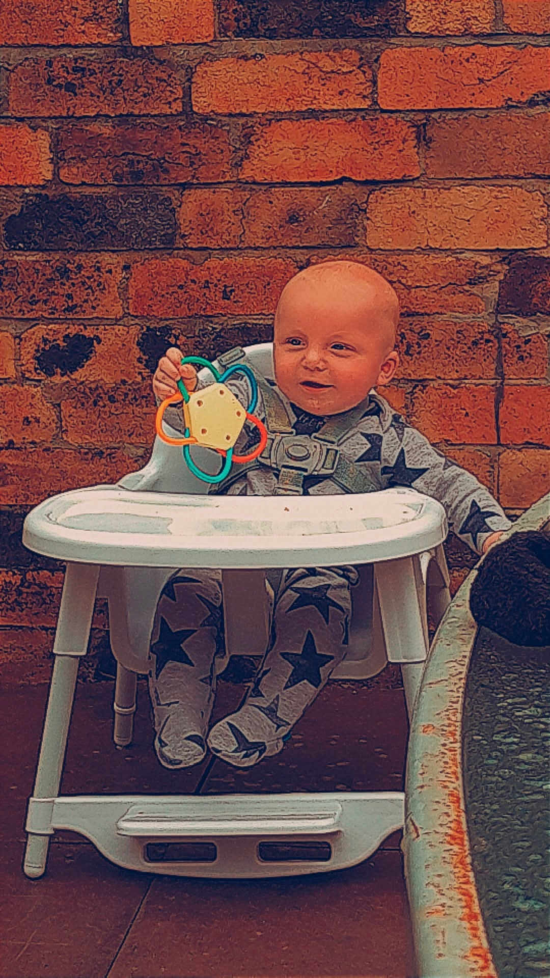 Leo is registered to the contest to win money with this photo: baby, high_chair, toy, brick_wall, pajamas, child, indoor, furniture, playful, smiling, seated, person, footwear, holding, colorful, face, expression, cute, young, infant