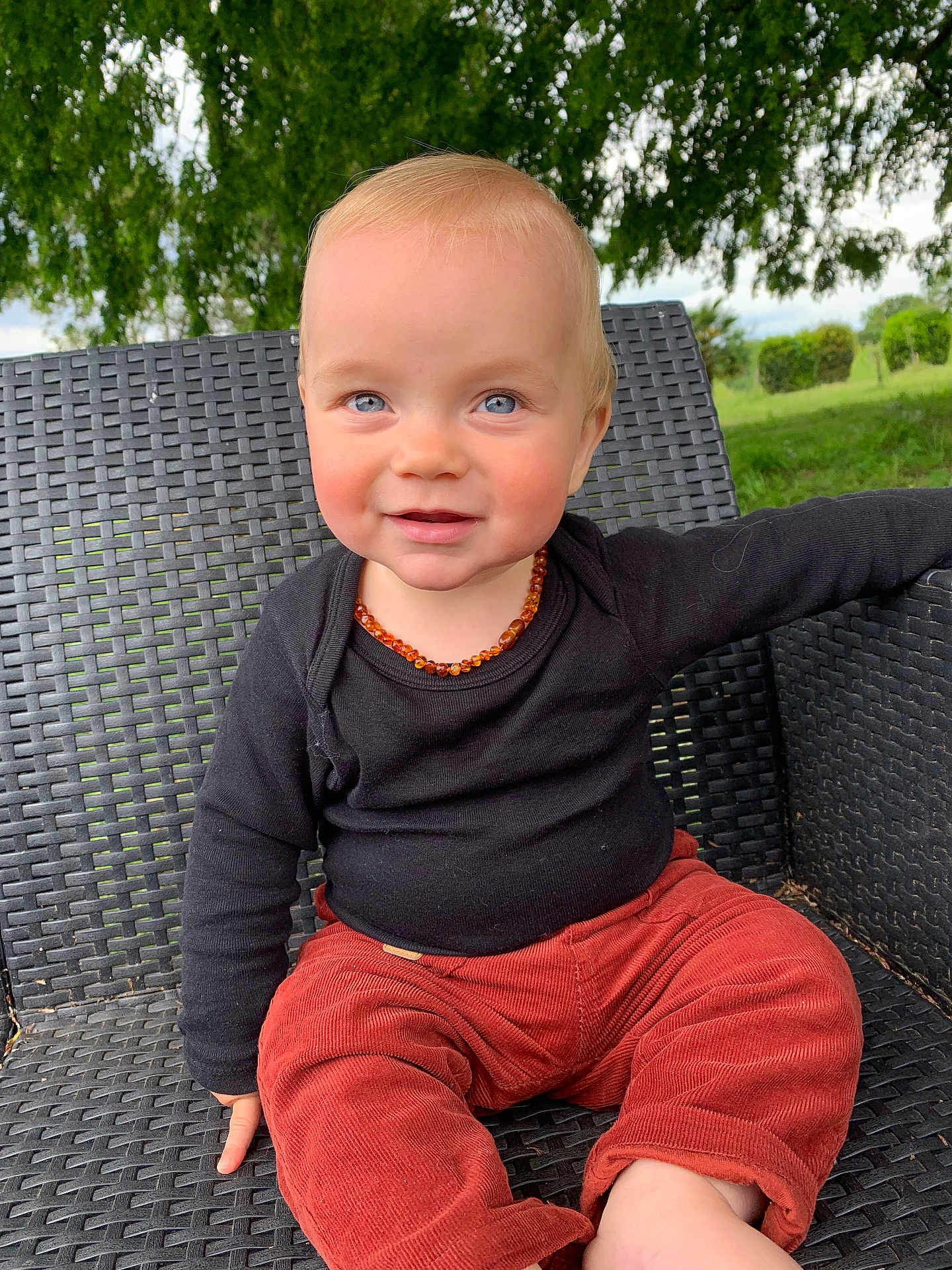 Mattìa a rejoint le concours — aidez-le/la à gagner de superbes lots ! toddler, child, smiling, outdoor, chair, woven_chair, black_shirt, red_pants, necklace, amber, blue_eyes, portrait, cute, happy, grass, trees, nature, casual_clothing, sitting, baby