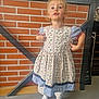 child, dress, patterned_clothing, socks, indoor, brick_wall, floor, metal_frame, playful_expression, standing, person, young_child, blonde_hair, long_sleeves, casual, cute, portrait, fashion, confident, home