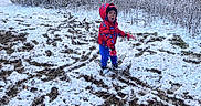 Aiden is registered to the contest to win money with this photo: blue_pants, child, clothing, cold_weather, field, footwear, frozen, gloves, hat, house, nature, outdoor, person, plants, red_jacket, snow, snowball, standing, surprised_expression, winter