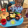 child, boy, mickey_mouse, table, food, plate, chair, cup, meal, shirt, indoor, floor, expression, happy, cute, kid, colorful, dining, snack, playful