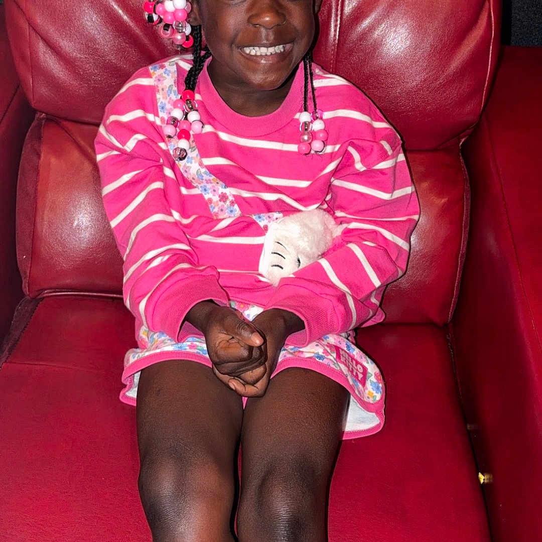 Harmoni joined the competition — help win amazing prizes! child, girl, smiling, braids, beads, pink_sweater, striped_clothing, floral_dress, red_leather_chair, seated, happy, indoor, portrait, young, fashion, cute, hands_folded, legs_stretched, casual, joyful
